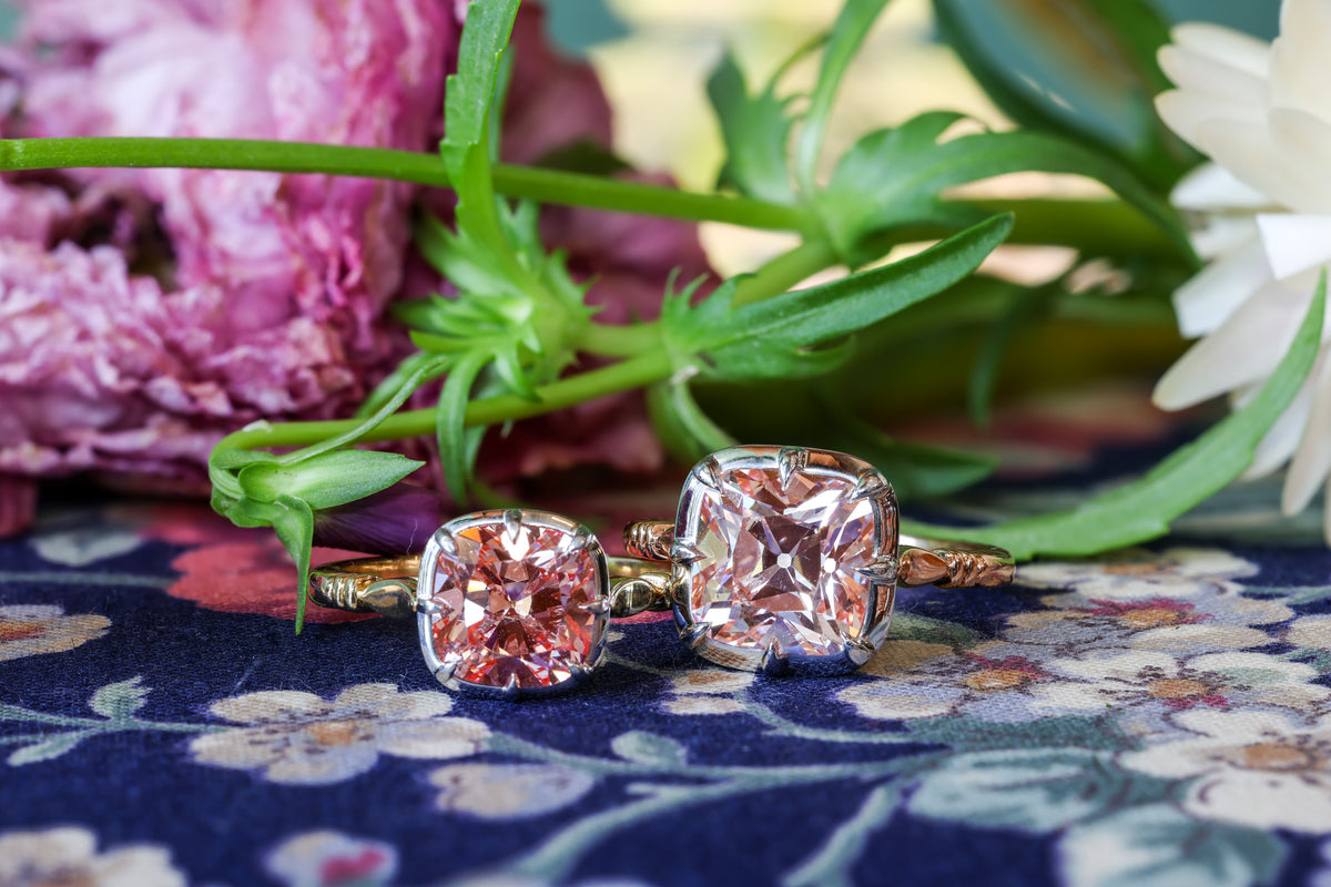 Two C. Disston collet rings with pink antique cushion-cut stones in yellow gold, resting on floral fabric among flowers